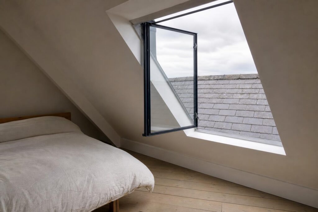 Side-hung black steel conservation rooflight swung open like a door low in a slate roof, seen from inside a loft bedroom
