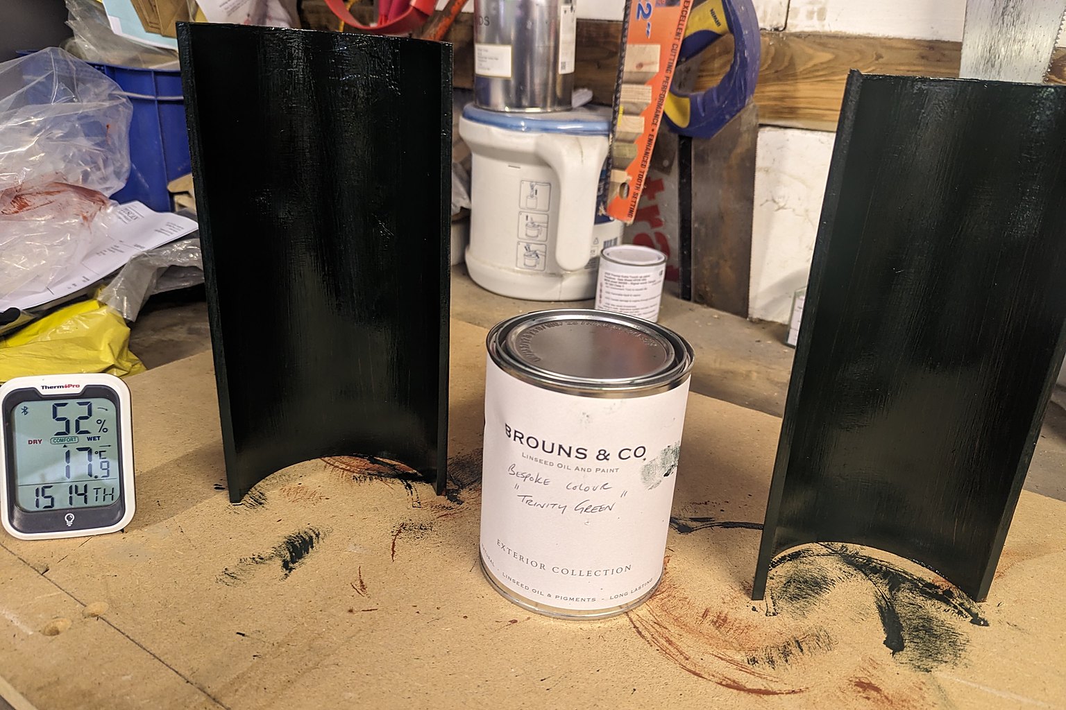 Two cast iron gutter sections freshly finished in a bespoke dark green linseed oil paint beside a Brouns and Co tin labelled Trinity Green, with a hygrometer on the workshop bench
