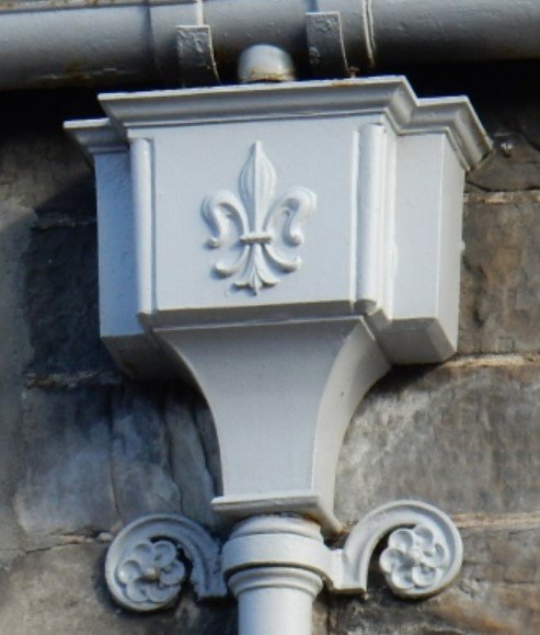 Cast iron hopper head with a fleur-de-lys motif, painted finish, installed on a stone building