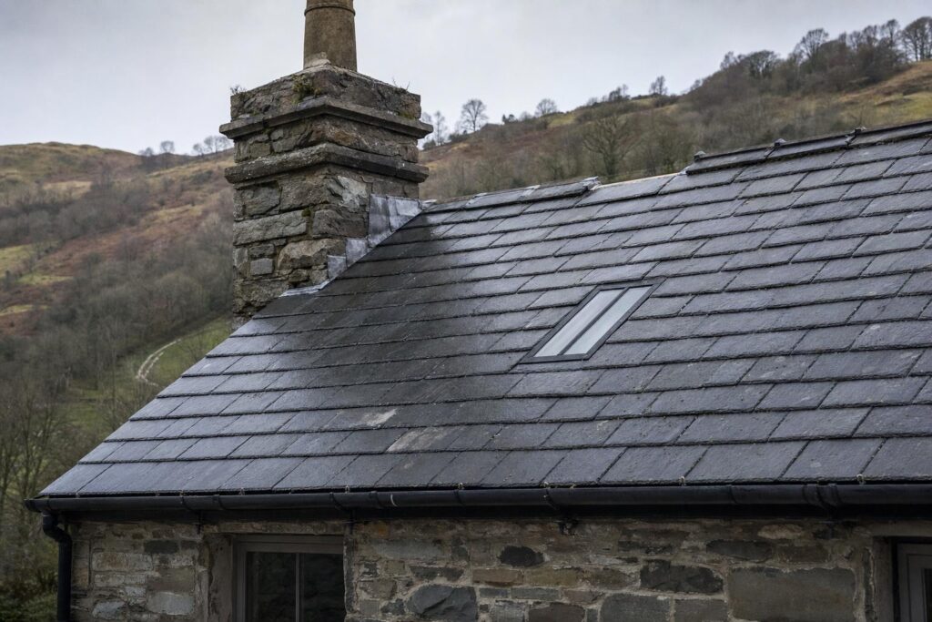 A small black steel conservation rooflight set flush into a weathered Welsh slate roof on a stone period building.