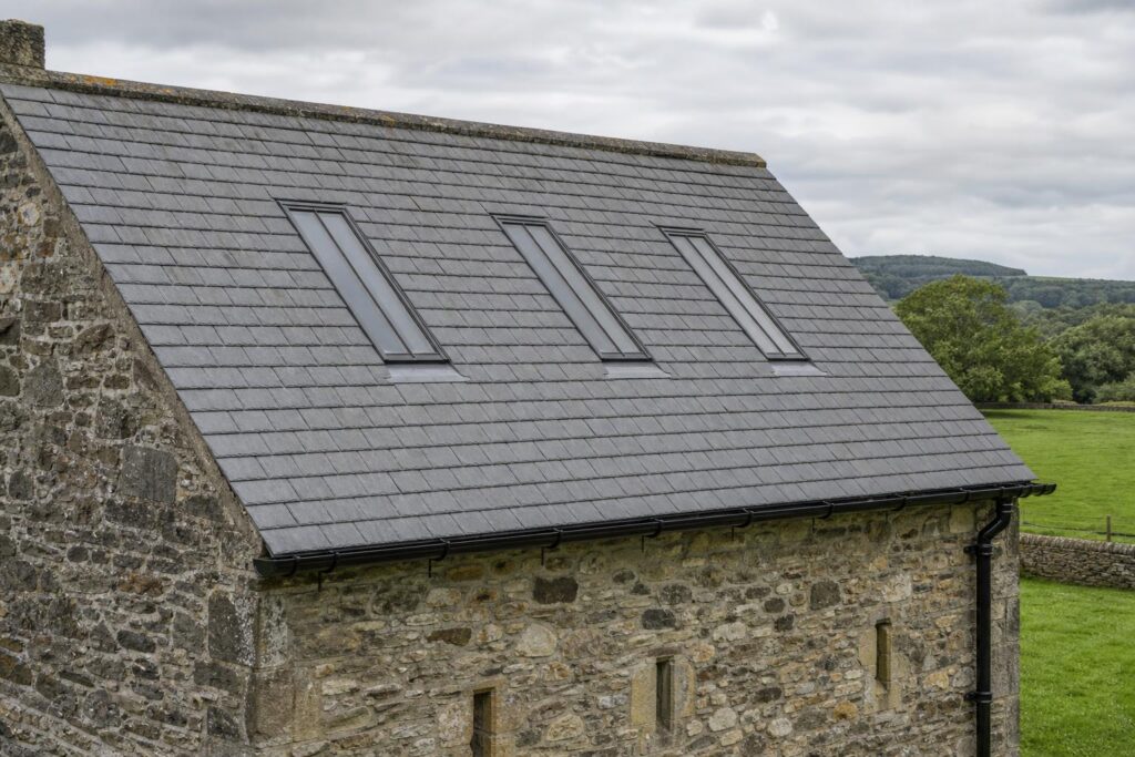 Three slim black steel conservation rooflights set flush in the slate roof of a stone barn