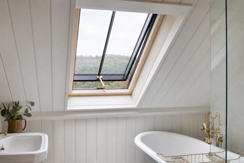 Slim black steel conservation rooflight open on a brass hand winder, seen from inside a bathroom with white-painted sloping ceiling