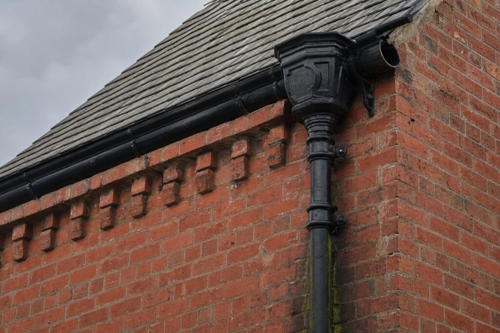 Black cast iron ogee gutter, moulded hopper head and downpipe on a Victorian red brick gable with a damp stain below a failed joint