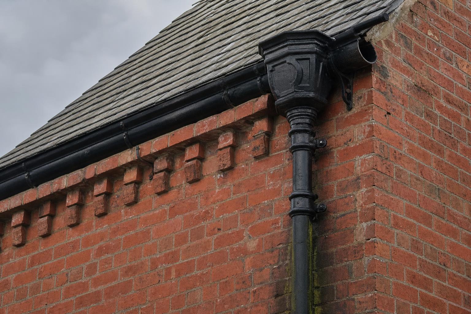 Black cast iron ogee gutter, moulded hopper head and downpipe on a Victorian red brick gable with a damp stain below a failed joint