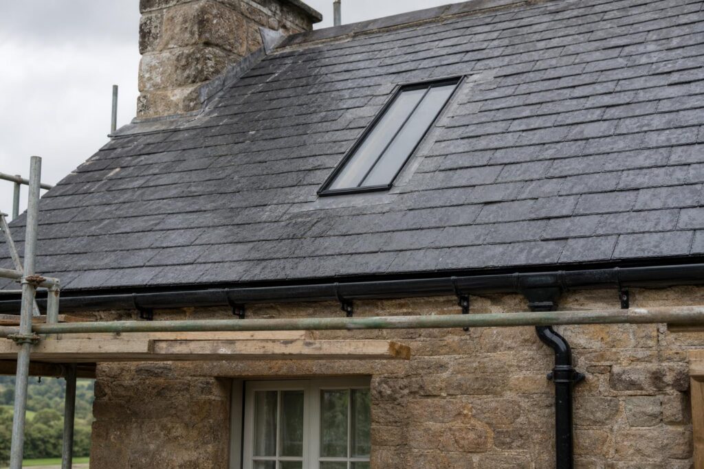 Scaffolded slate roof on a stone building with a slim black steel conservation rooflight and black cast iron gutters at the eaves