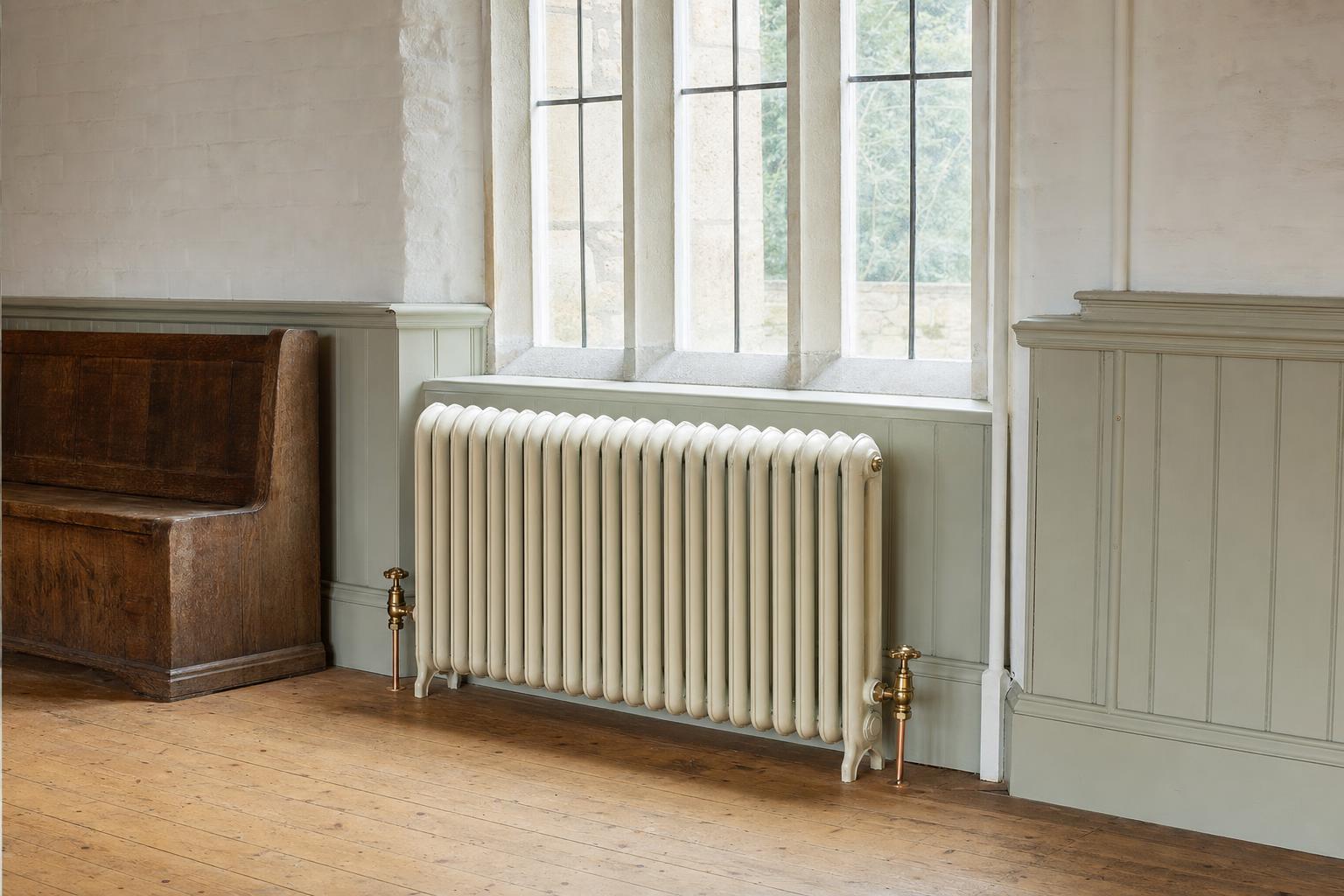 Board School two-column cast iron radiator with rounded tops beneath a tall window in a Victorian village hall.