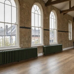 Board School three-column cast iron radiators with rounded tops beneath tall windows in a converted Victorian schoolroom.