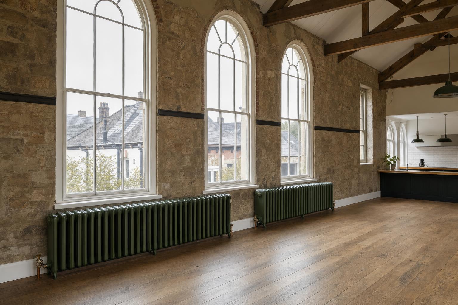 Board School three-column cast iron radiators with rounded tops beneath tall windows in a converted Victorian schoolroom.