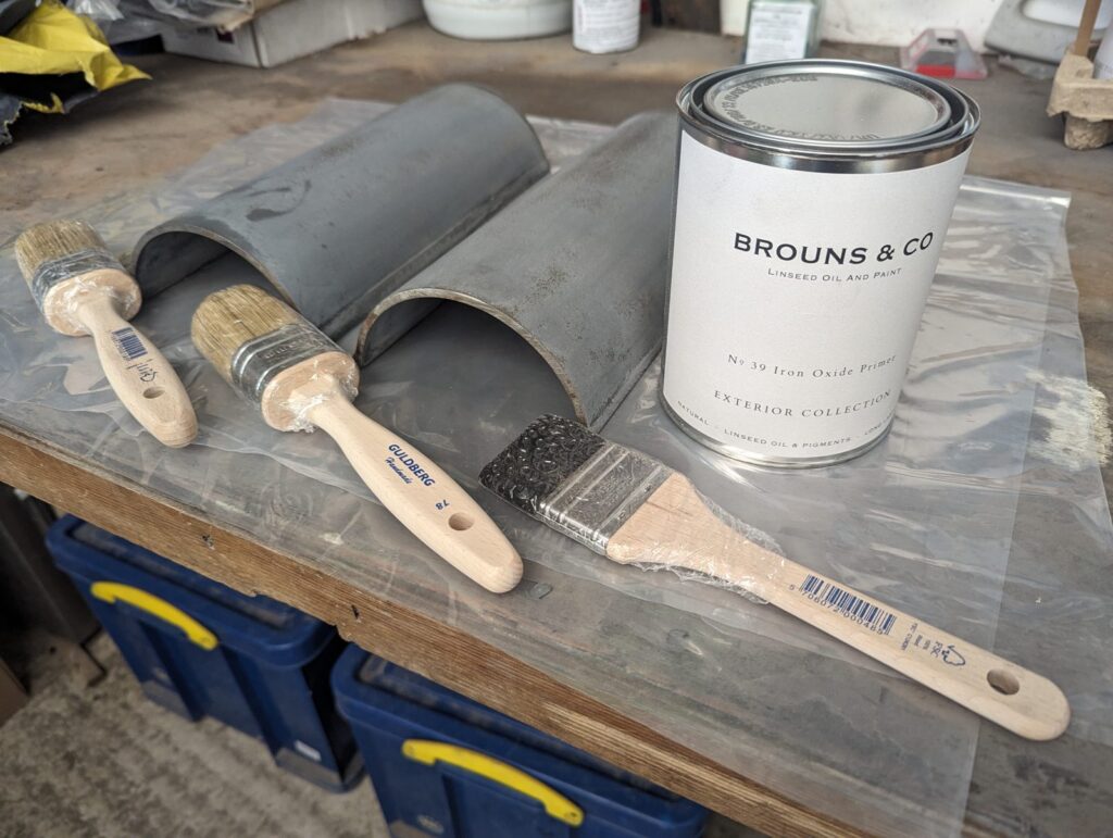 Primed cast iron gutter sections on a bench with a tin of linseed oil paint and brushes