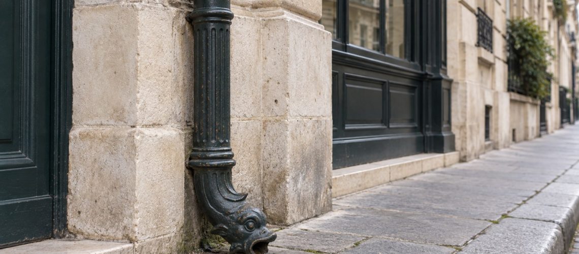 Fluted cast iron dauphin with fish-head spout at the foot of a zinc downpipe on a Parisian limestone façade