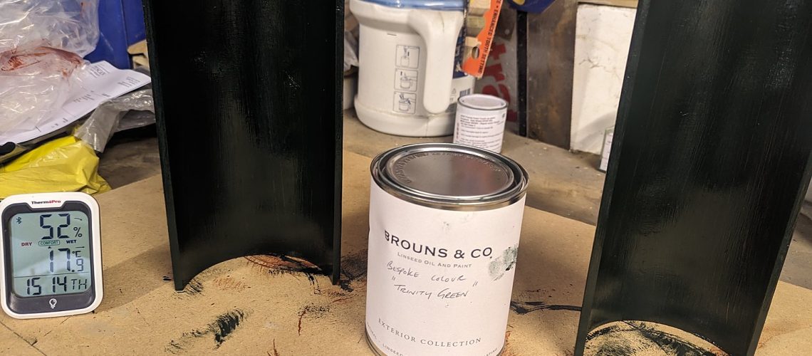 Two cast iron gutter sections freshly finished in a bespoke dark green linseed oil paint beside a Brouns and Co tin labelled Trinity Green, with a hygrometer on the workshop bench