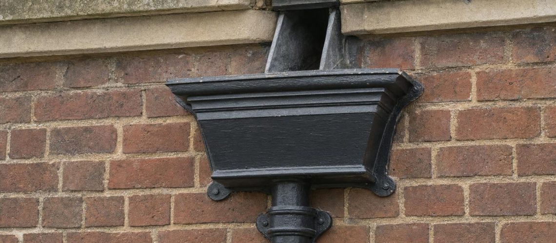 An ornamental cast iron hopper head collecting water from a parapet outlet above a round cast iron downpipe on a Georgian brick wall.