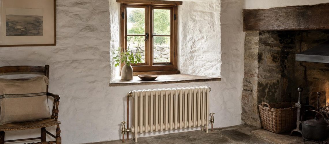 Low cream cast iron column radiator beneath a deep-set window in a thick-walled stone cottage.