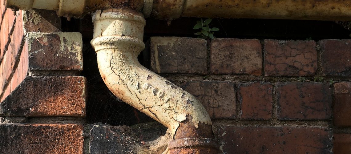 Cast iron half round gutter and downpipe offset on a red brick wall with rust breaking through the paint, flaking finish and a cracked socket joint, showing typical signs of failing cast iron guttering